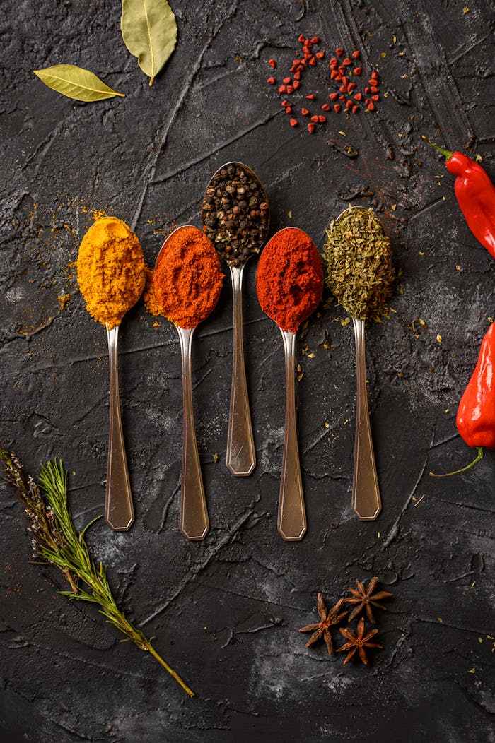 services-04 Vibrant spices displayed on spoons with chili peppers on a dark textured surface, emphasizing culinary art.